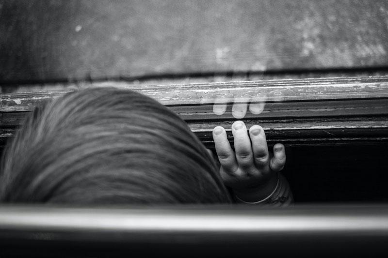 Black and white photo of the top of a child's head with their hand resting on a window.