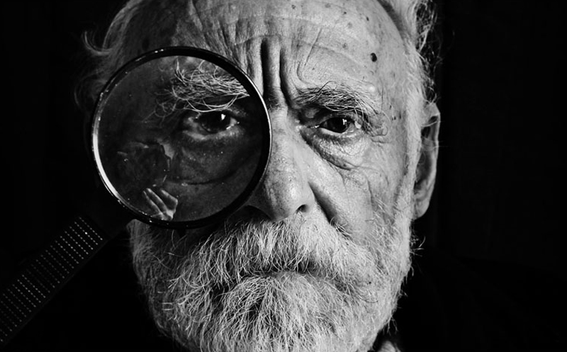 Black and white photo of an older man holding a maginifying glass to his right eye.