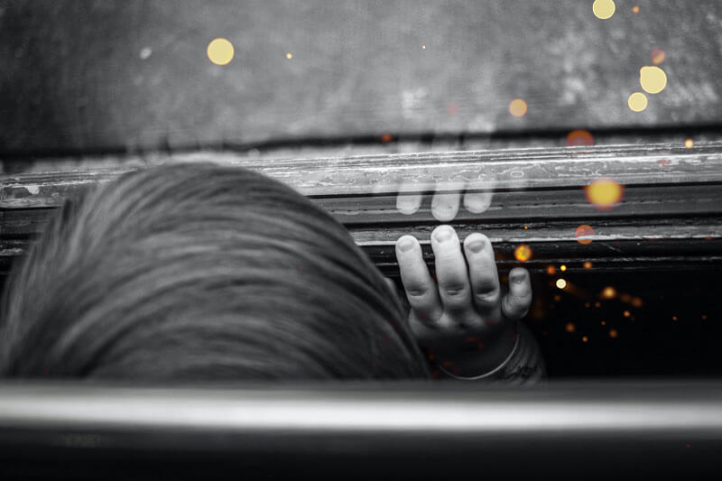 Black and white photo of the top of a child's head with their hand resting on a window.