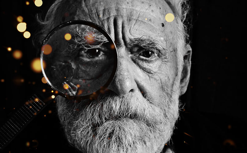 Black and white photo of an older man holding a maginifying glass to his right eye.